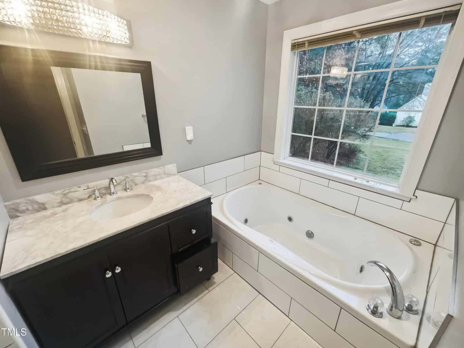 Bathroom with a large rectangular bathtub and a dark wood vanity. The vanity has a marble top, a sink with a silver faucet, and two open drawers. Above is a mirror with a light fixture. A window with blinds is in front of the bathtub.