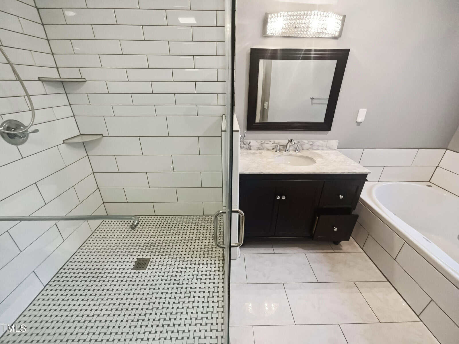 Modern bathroom with a glass-enclosed shower featuring white subway tiles and a small hexagonal floor pattern. A dark wood vanity with a marble countertop and rectangular mirror is adjacent to a white bathtub. The lighting fixture is above the mirror.