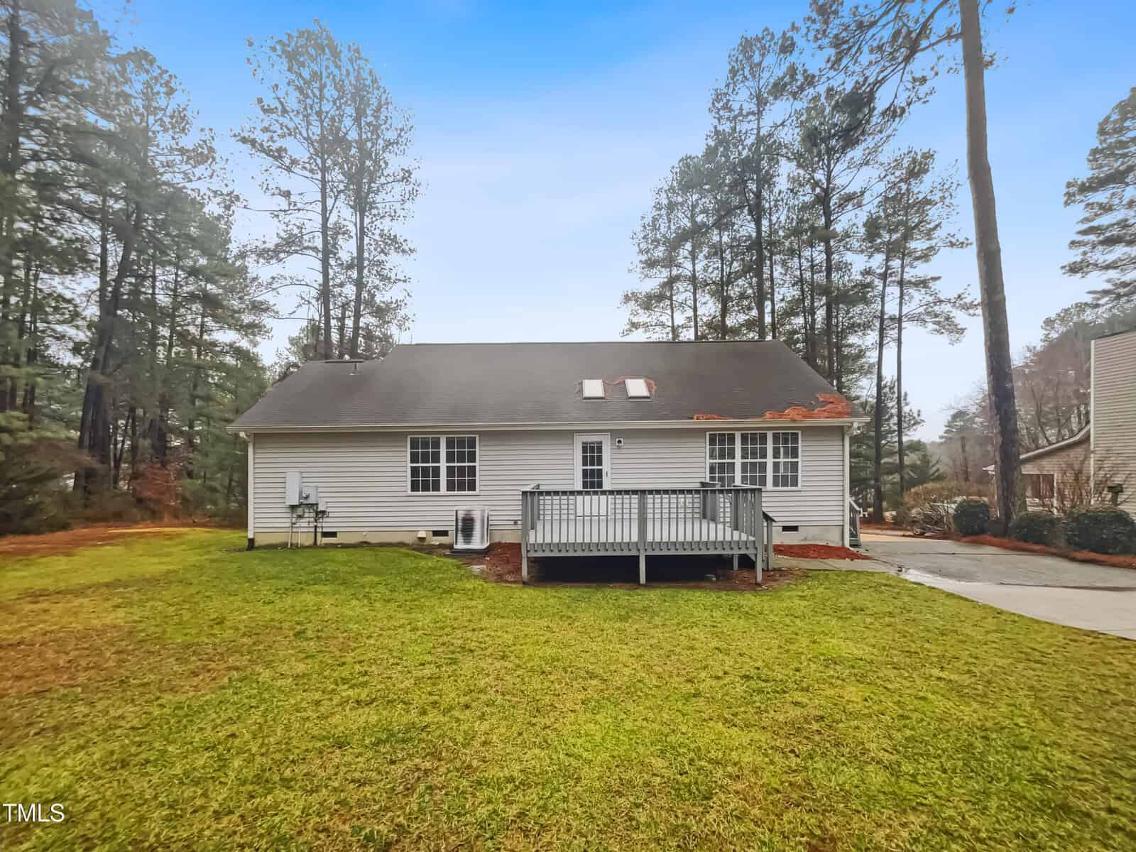 A one-story house with white siding, two skylights on the roof, and a small wooden deck at the back. The house is surrounded by a well-maintained lawn and tall pine trees, under a clear blue sky. A driveway is visible on the right.