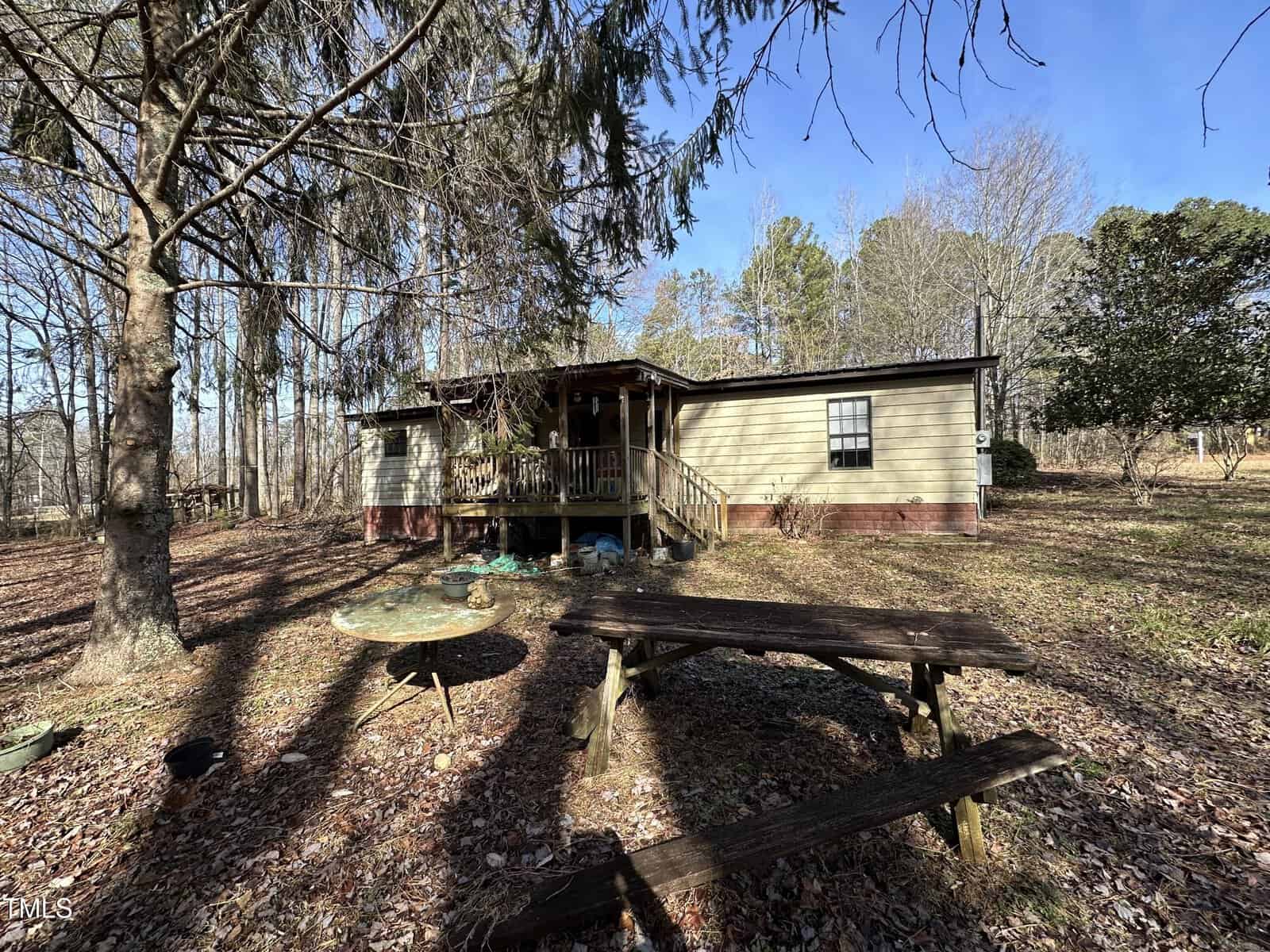 A single-story house with a covered front porch sits in a yard with fallen leaves. In the foreground are a weathered wooden picnic table and a round glass table. Leafless trees and some evergreens surround the home.