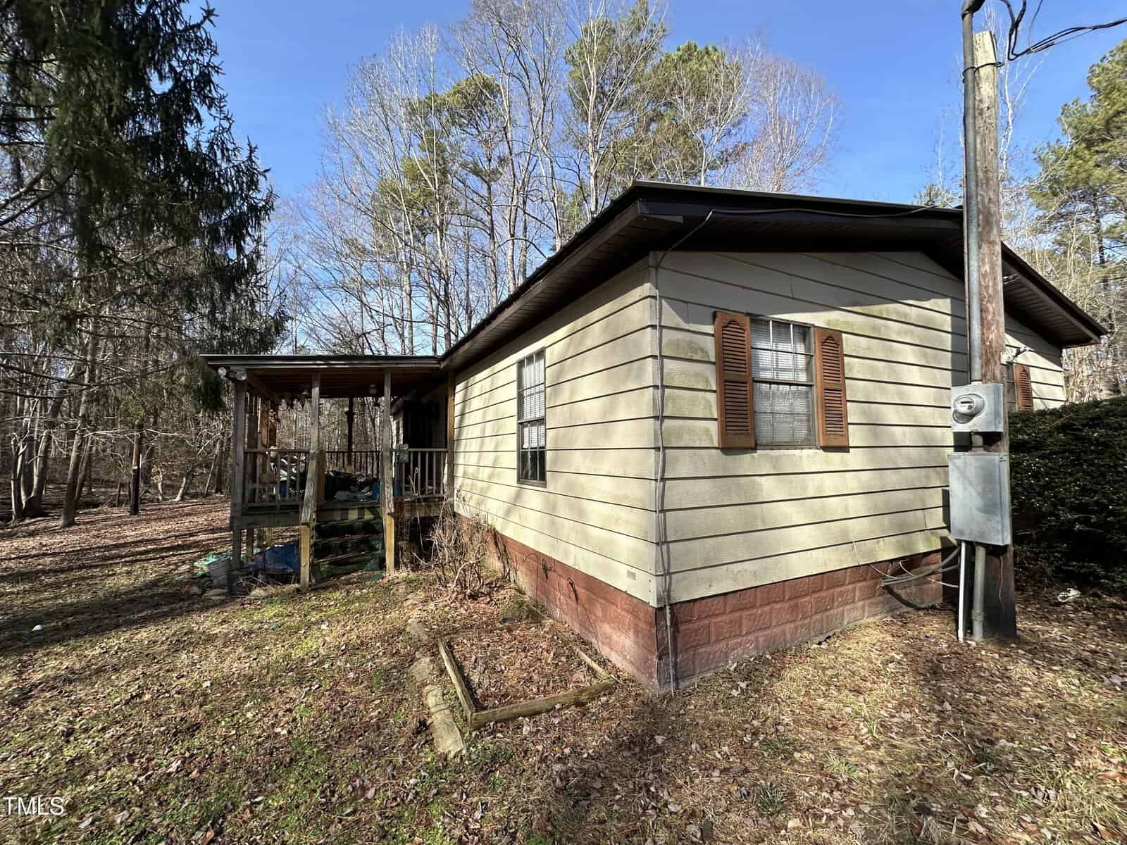 A single-story house with beige siding, brown shutters, and a brick foundation sits on a wooded lot with a covered porch and visible utility meter. Leafy ground and bare trees surround the house.