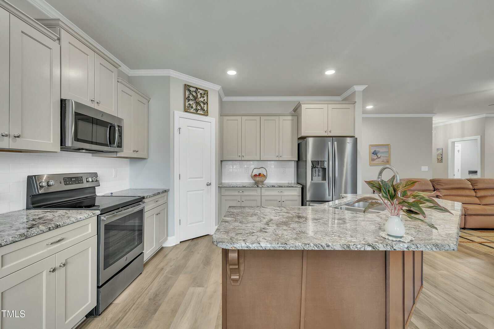 Modern kitchen with light gray cabinets, stainless steel appliances, a center island with granite countertop, a plant in a pot, wood flooring, and recessed lighting. Refrigerator and oven are visible. Open layout leads to living area.
