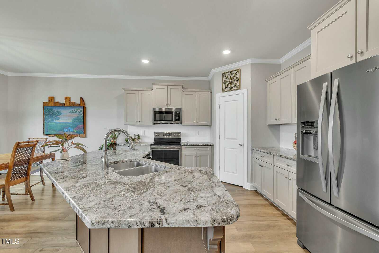 Modern kitchen with light gray cabinets, stainless steel appliances, marble-patterned countertops, and a kitchen island with a sink. Dining area with a table and chairs is visible in the background. Light wood flooring throughout.