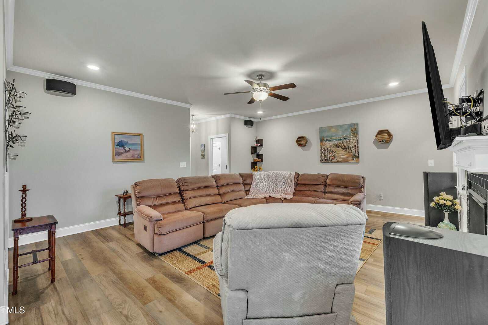 A living room with beige walls, wood flooring, a large sectional sofa, a recliner, wall art, a ceiling fan, a wall-mounted TV, and a fireplace. A small table and a carpet are also visible.