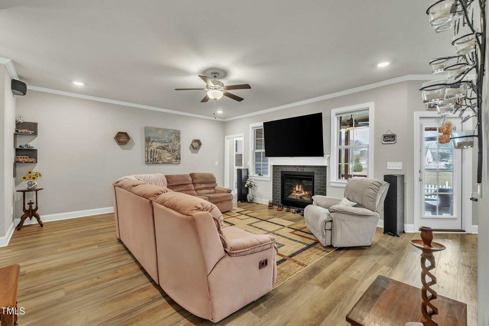 A living room with beige reclining sofas, a matching armchair, a fireplace with a mounted TV above it, wood flooring, wall shelves, and windows providing natural light. A ceiling fan and artwork complete the decor.