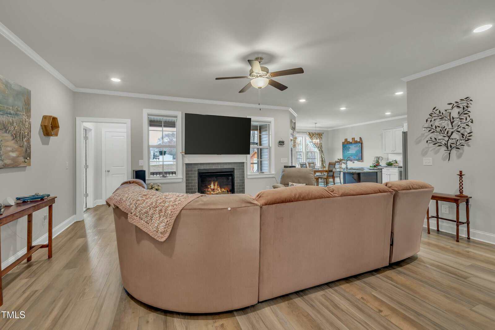 A living room with a curved beige sectional sofa facing a wall-mounted TV above a fireplace, light wood floors, ceiling fan, and an open view into the kitchen and dining area.