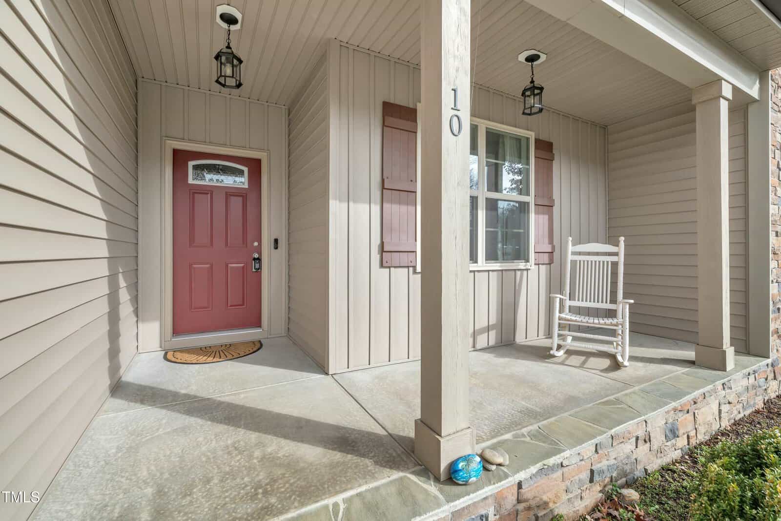 A covered porch with beige siding features a red front door, two hanging light fixtures, a white rocking chair, and the number 10 on a column. The porch floor is concrete, and there is a small welcome mat by the door.