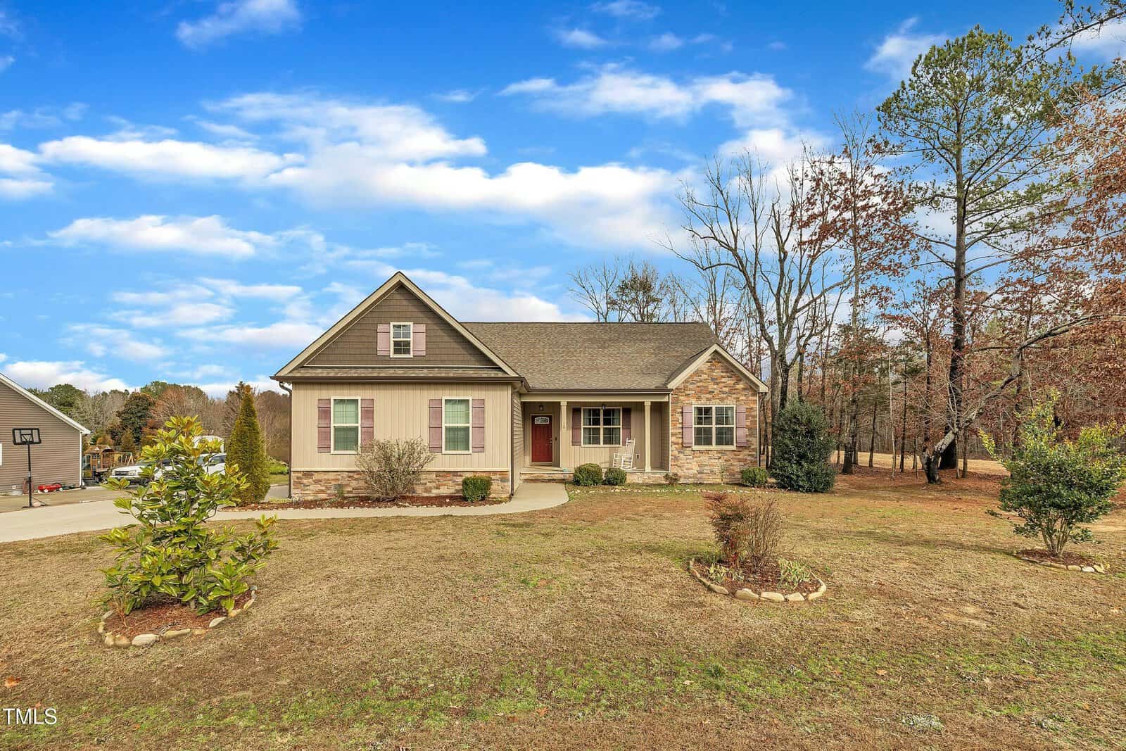A single-story house with beige siding, stone accents, and a gabled roof. It features a red front door, a small covered porch, and a landscaped front yard with a curved driveway and sparse trees in the background.