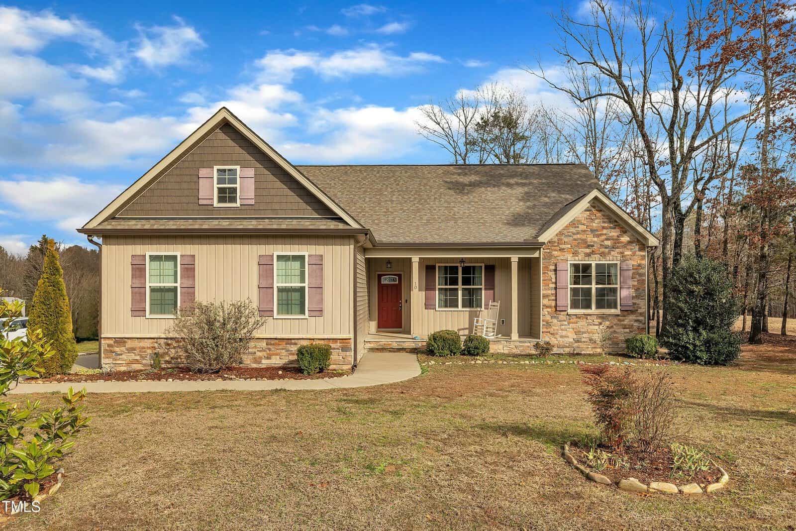 Single-story house with tan siding, stone accents, and brown shutters, located on a spacious, partially landscaped yard with a concrete walkway leading to the front door and rocking chair on the porch. Trees border the property.