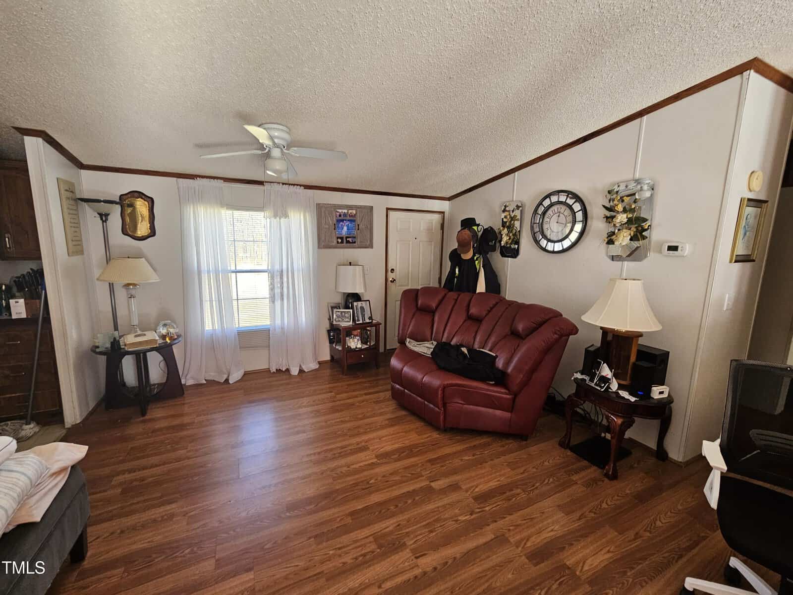 A living room with a red leather sofa, wooden flooring, and a ceiling fan. Walls are adorned with a clock and decorative items. A window with sheer curtains is on the left, and a side table with a lamp is nearby. A coat rack stands by the door.