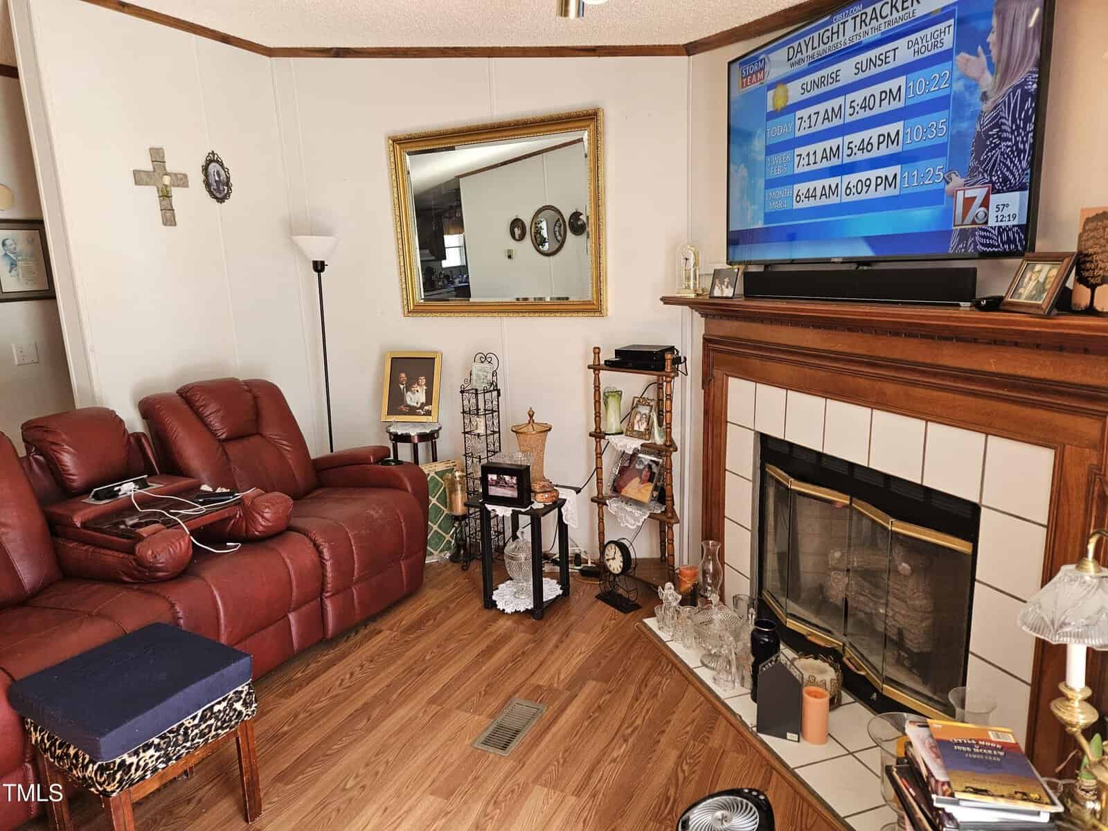 A living room with a brown leather recliner, a wooden floor, and a fireplace adorned with decorative items. Above the fireplace is a TV displaying a weather channel. A large mirror hangs on the wall, and various photos and knickknacks are visible.