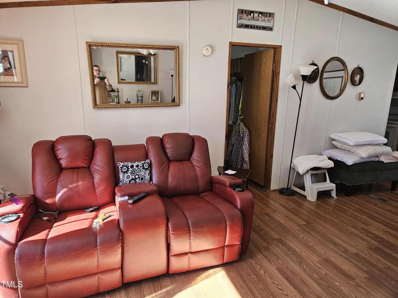 A living room with two red recliner chairs on a wooden floor. A mirror and framed pictures hang on the wall. A lamp stands next to a closet with clothes. A small set of shelves holds folded linens, and circular mirrors hang above it.