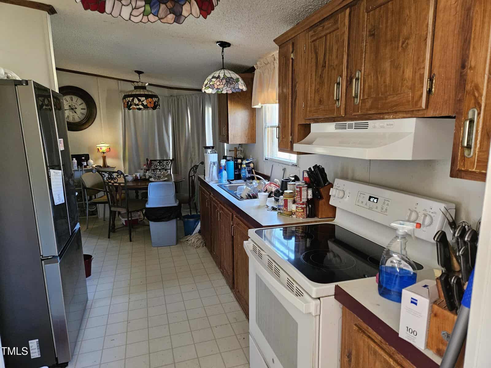 A kitchen with a white stove and wooden cabinets. The counter is cluttered with various items, including a blue spray bottle and canned goods. A table with chairs is visible in the background, alongside a clock on the wall and hanging lights.