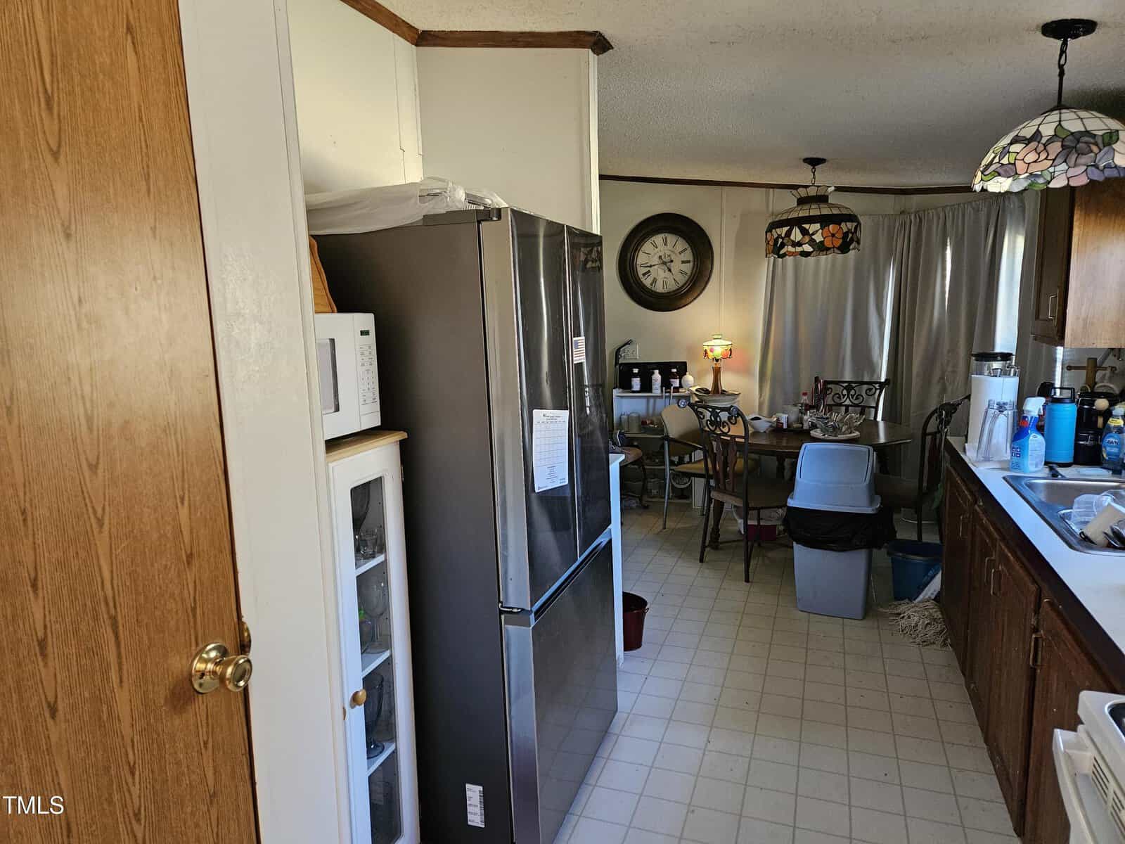 A kitchen with a stainless steel refrigerator, wooden cabinets, and a countertop with various items. A microwave sits on a small cabinet. A dining table with chairs is visible in the background. The floor is tiled, and stained glass lamps hang from the ceiling.