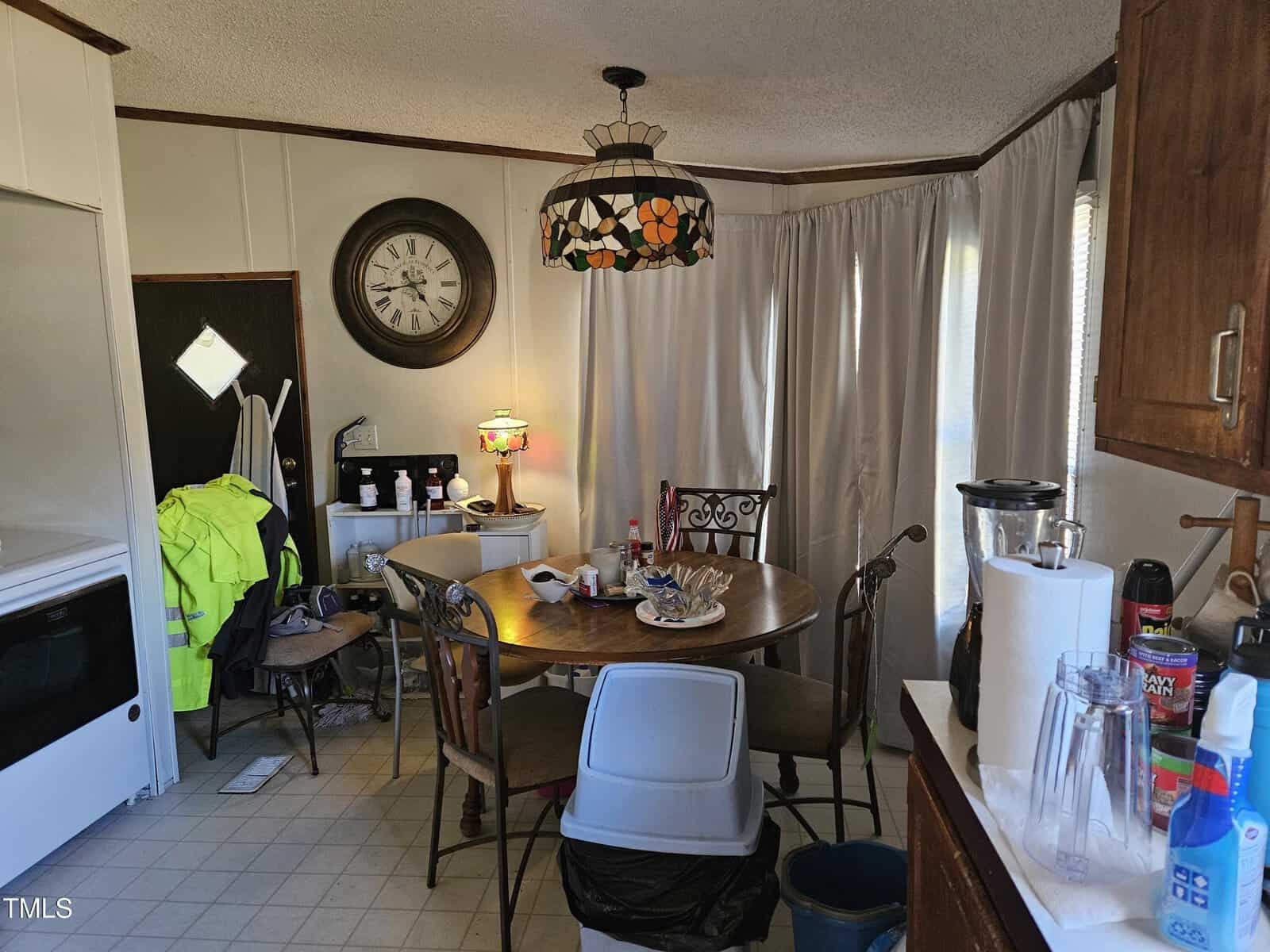 A kitchen with a round table and four chairs. A decorative light hangs above. The table holds miscellaneous items and a cooler. A large wall clock is visible. A high-visibility jacket hangs on a nearby door. Appliances and curtains are in view.