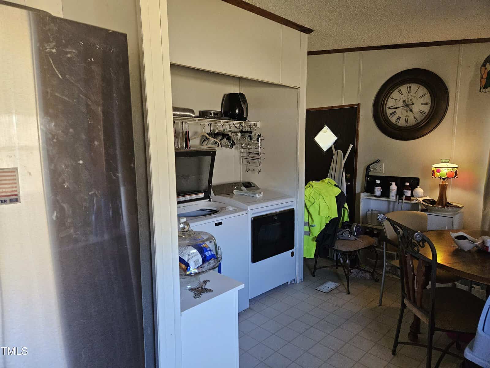 A cluttered laundry room with a washer and dryer set, shelves holding various items, a large wall clock, a dining table with chairs, and a lamp on the table. A high-visibility jacket hangs nearby.