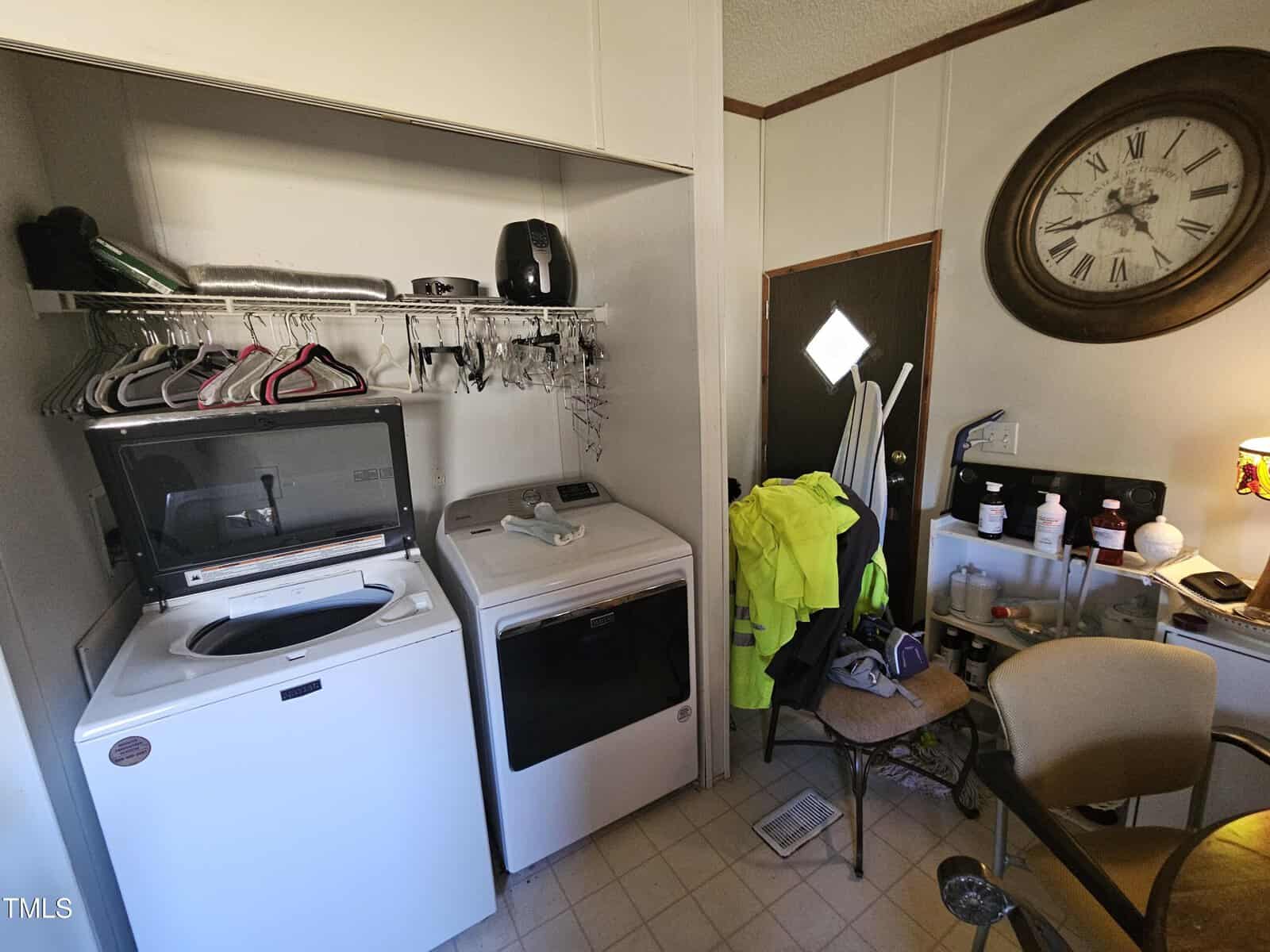 A laundry room with a washing machine and dryer set side by side. A shelf above holds hangers, detergent, and a black appliance. A large wall clock and bright yellow jackets hang on the far wall. An office chair and desk are on the right.