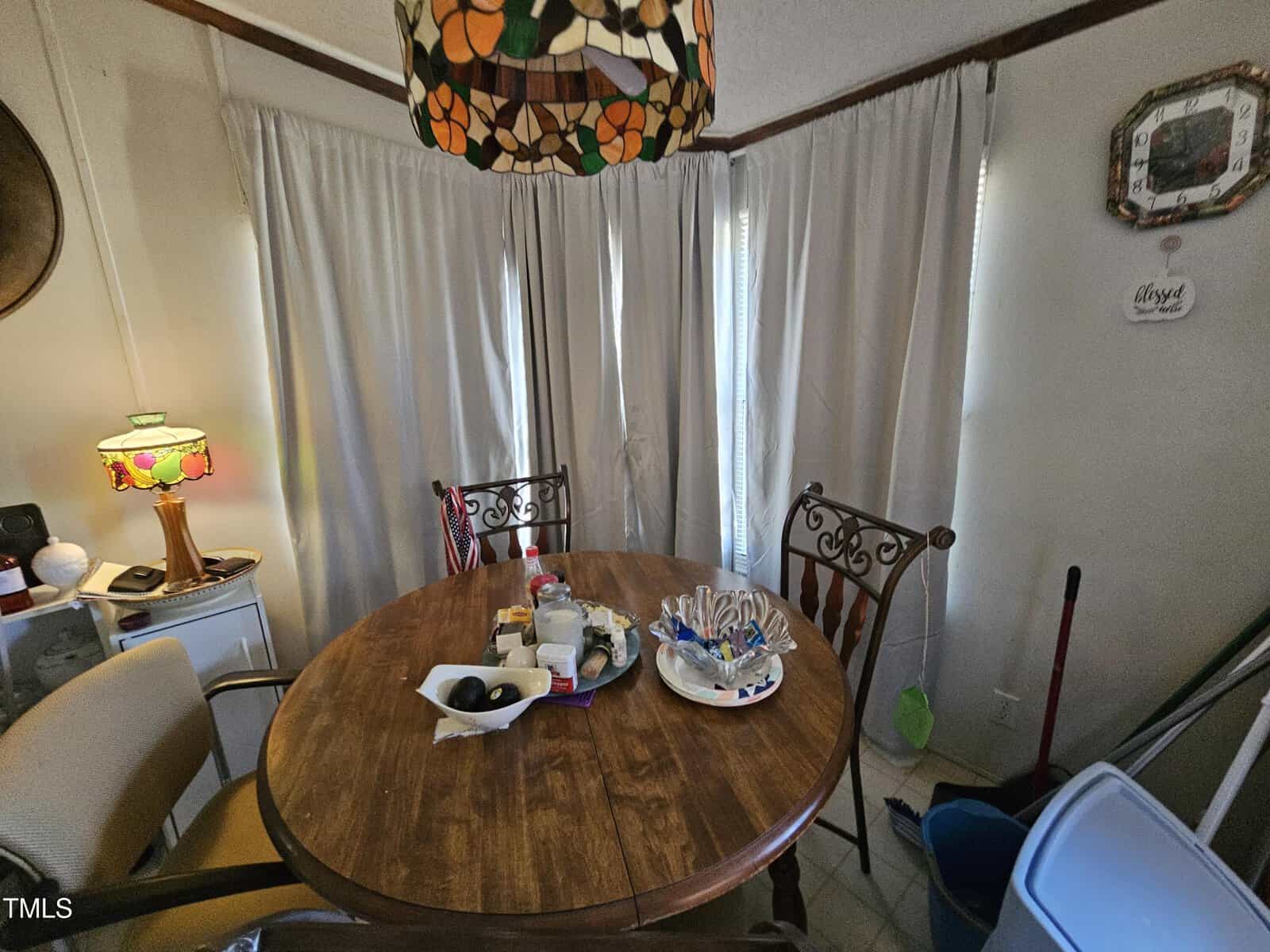 A dining area with a round wooden table surrounded by four chairs. The table is cluttered with various items like bottles and a basket of utensils. A colorful lamp is on a stand in the corner. Beige curtains cover the windows in the background.