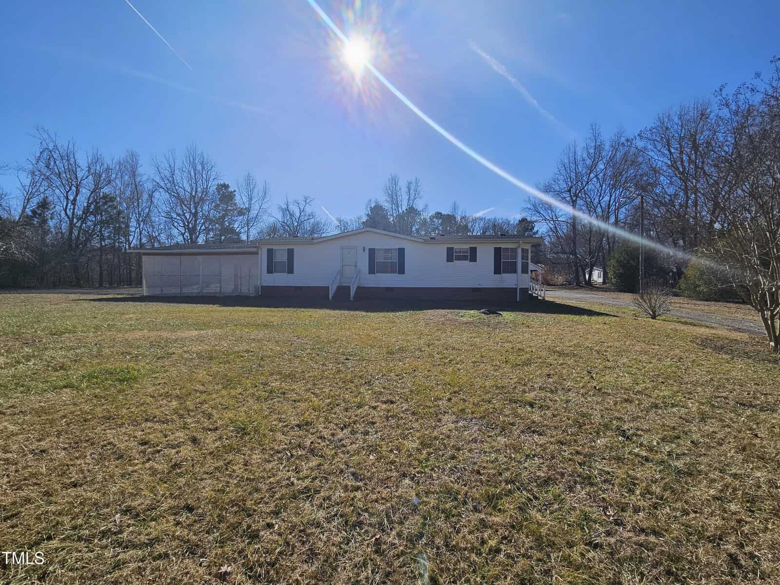 A single-story mobile home rests peacefully at 2020 Sanders Road in the middle of a grassy field under a clear blue sky. The sunlight is bright, casting shadows on the house and lawn. Bare trees surround the field like a natural stem, indicating a winter or early spring setting.