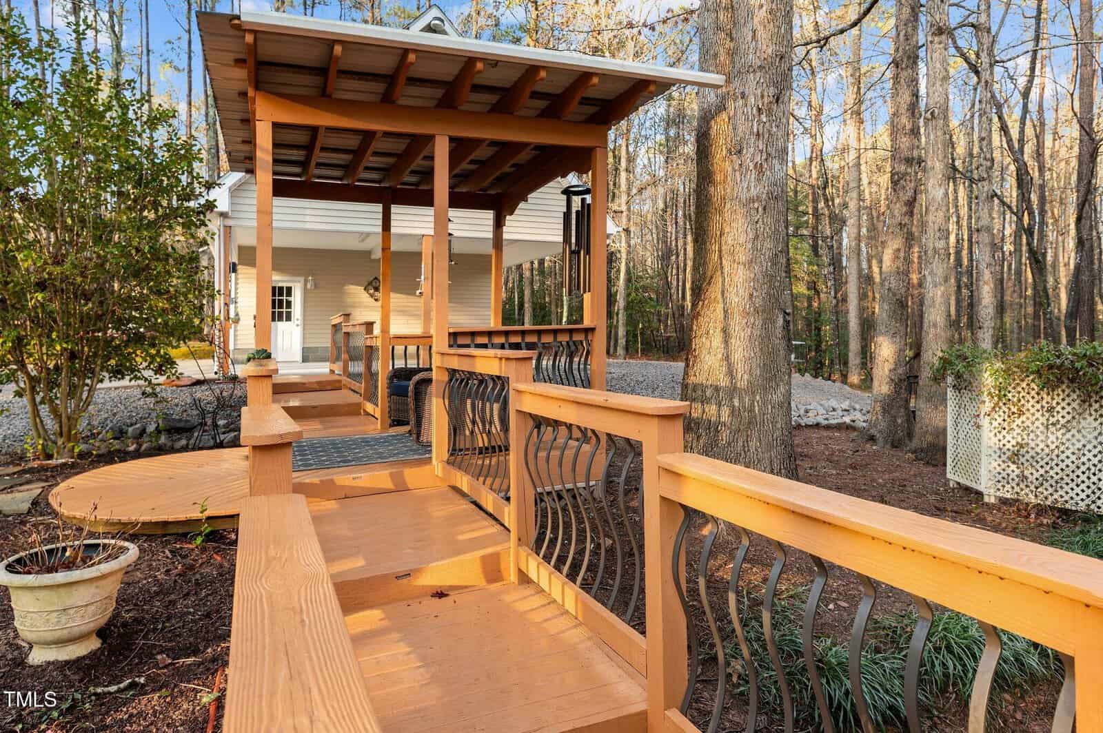 Wooden ramp with metal railings leads to a covered entrance of a white house surrounded by trees and landscaping, with a potted plant and trellis visible nearby.