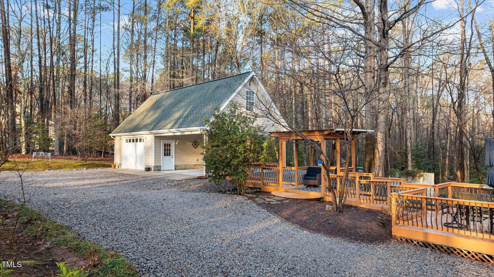 A light-colored two-story garage with a pitched roof sits next to a gravel driveway. A wooden gazebo structure and deck are on the right, surrounded by tall trees and natural landscaping.