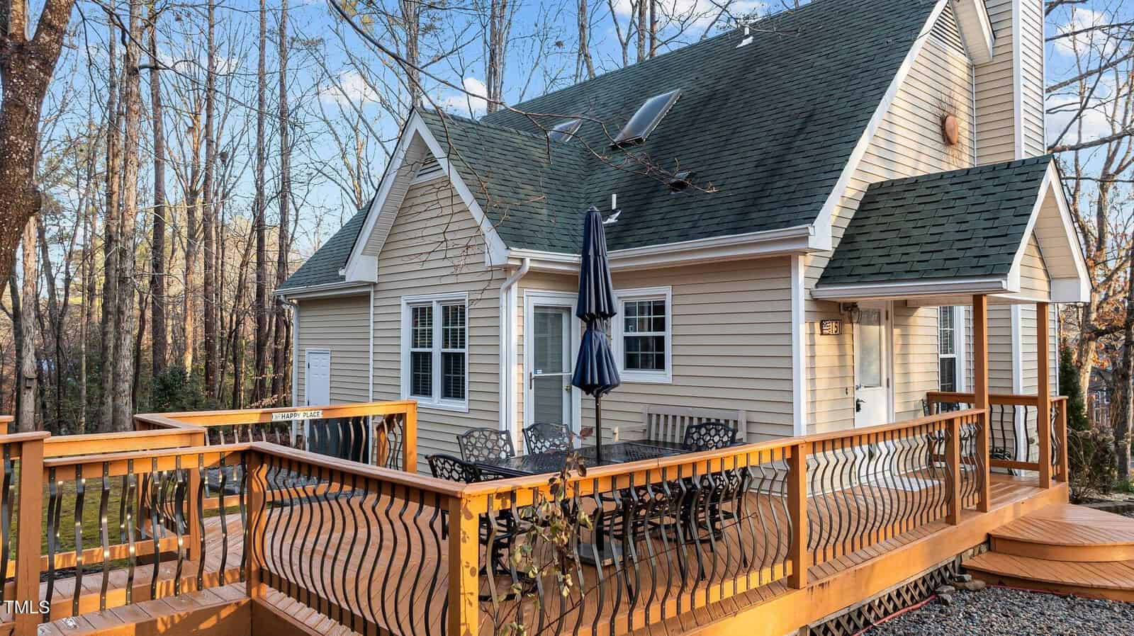 A tan house with green shingles features a large wooden deck. The deck has a metal outdoor dining set with an umbrella. Trees without leaves surround the house, and sunlight filters through the branches.