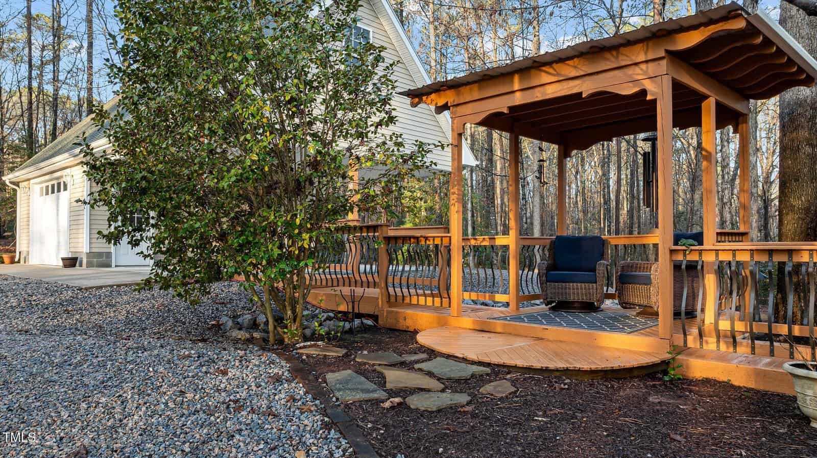 A wooden gazebo with seating stands on a deck near a gravel path and stepping stones, adjacent to a house with a white garage. Trees without leaves surround the area.
