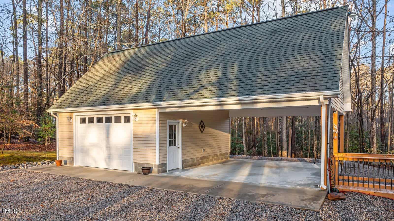 A detached garage with light-colored siding, a single garage door, a side entrance door, and a covered carport area sits on a gravel driveway surrounded by trees.