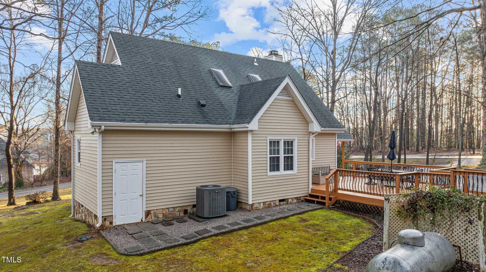 A beige house with dark gray shingles features two skylights, multiple windows, an outdoor HVAC unit, and a wooden deck with railings. The yard has sparse grass, trees, and a large propane tank in the foreground.