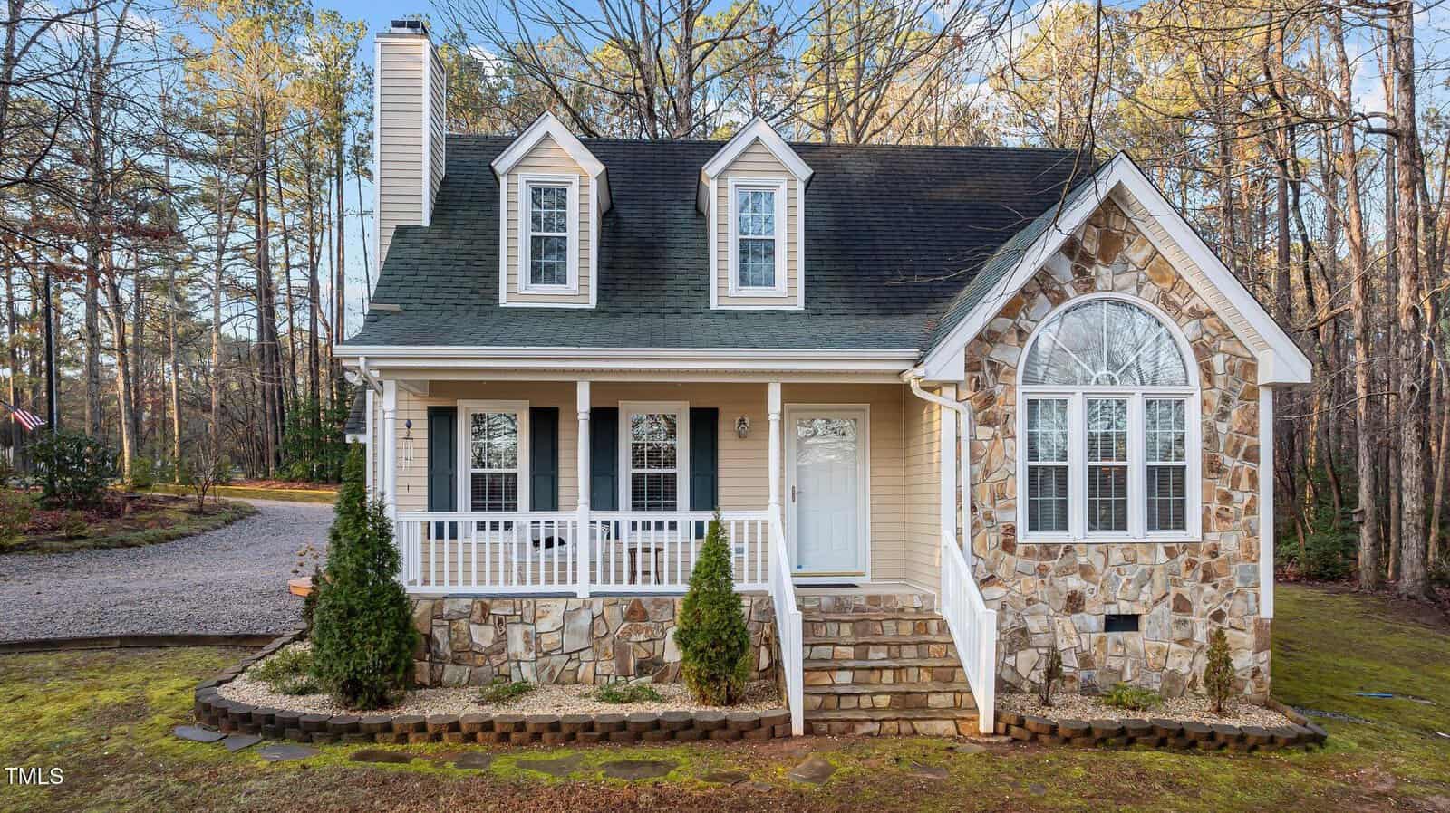 A small two-story house with a dark roof, stone and siding exterior, front porch with white railings, and an arched window, surrounded by trees and a gravel driveway.