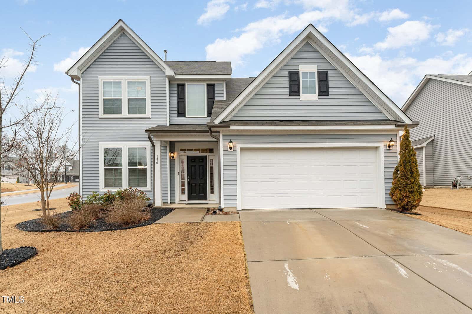 Two-story gray house with white trim, black shutters, and a double garage. The driveway and front yard show well-maintained landscaping with bushes and a small tree. The sky is partly cloudy.
