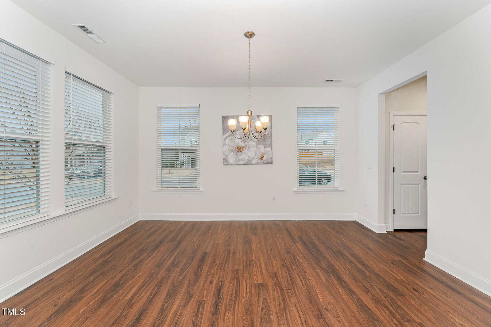 Empty dining room with three windows, a hanging light fixture, white walls, wood-look flooring, a floral wall art piece, and a white door leading to another room. Natural light enters through the windows.