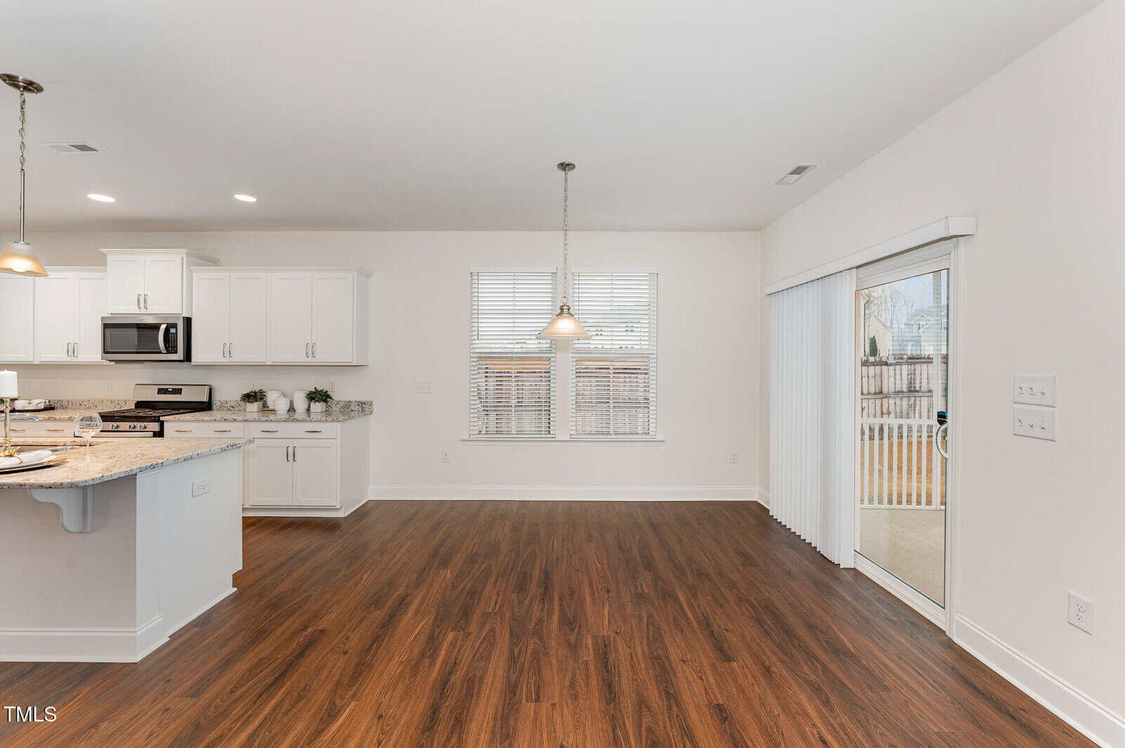 A modern kitchen and dining area with white cabinets, granite countertops, pendant lighting, wood-look flooring, and a sliding glass door leading to a patio. Large windows provide natural light.