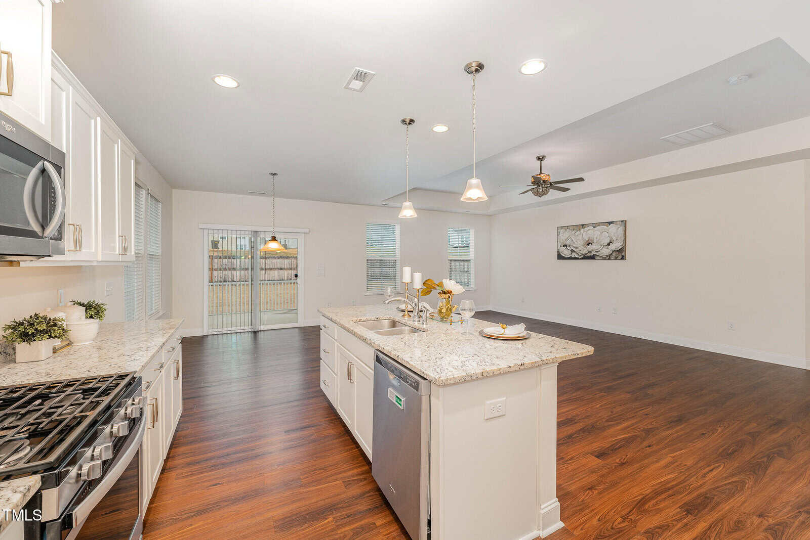 Open-plan kitchen and living area with white cabinets, granite countertops, stainless steel appliances, pendant lighting, hardwood floors, and sliding glass doors leading outside. Dining and living spaces are adjacent to the kitchen.