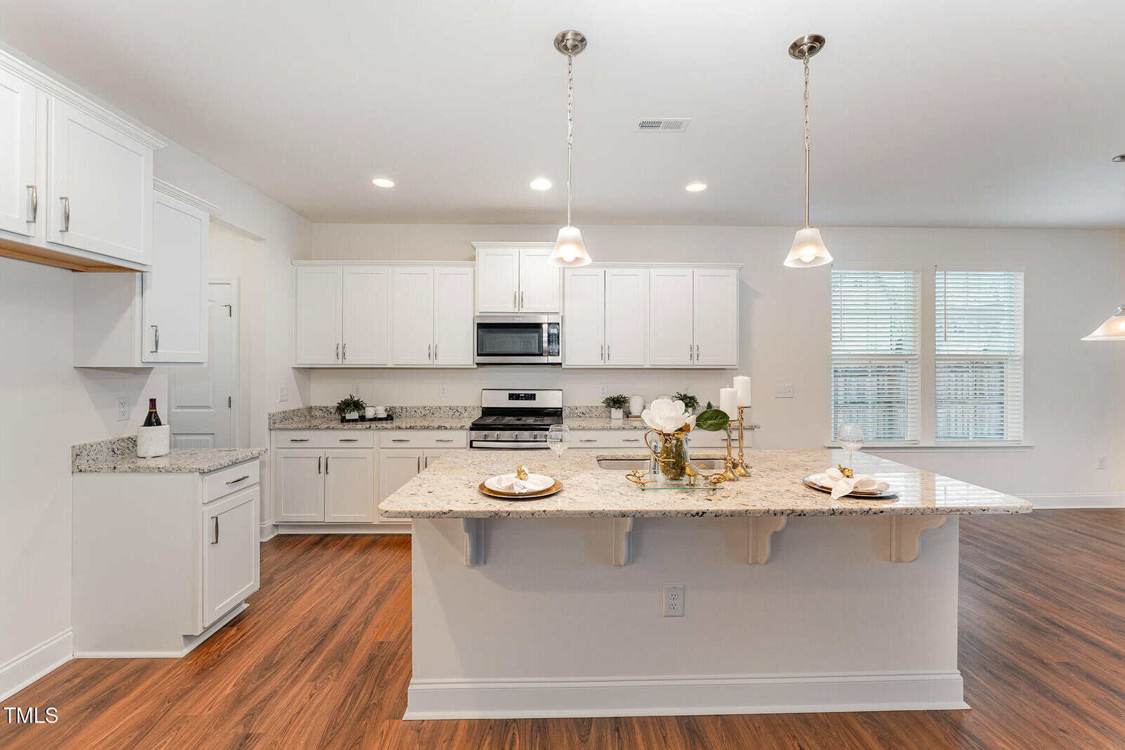 Modern kitchen with white cabinets, granite countertops, stainless steel appliances, and a large island with two place settings. Pendant lights hang above the island. The floor is medium-tone wood, and windows let in natural light.
