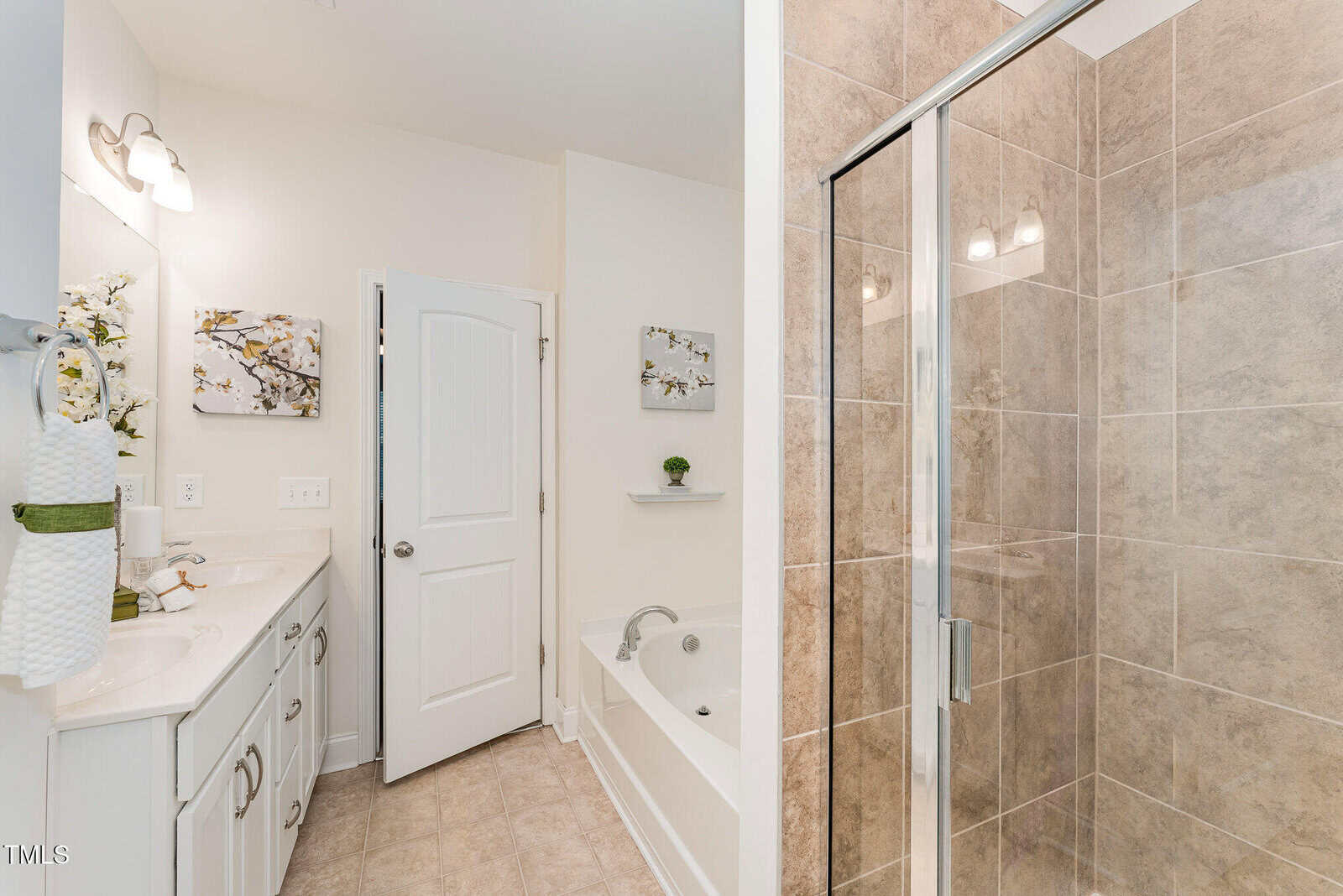 Modern bathroom with a glass-enclosed tiled shower, white bathtub, double vanity with sinks, large wall mirror, and neutral decor on the light-colored walls. Towels and flower arrangements are on the vanity.