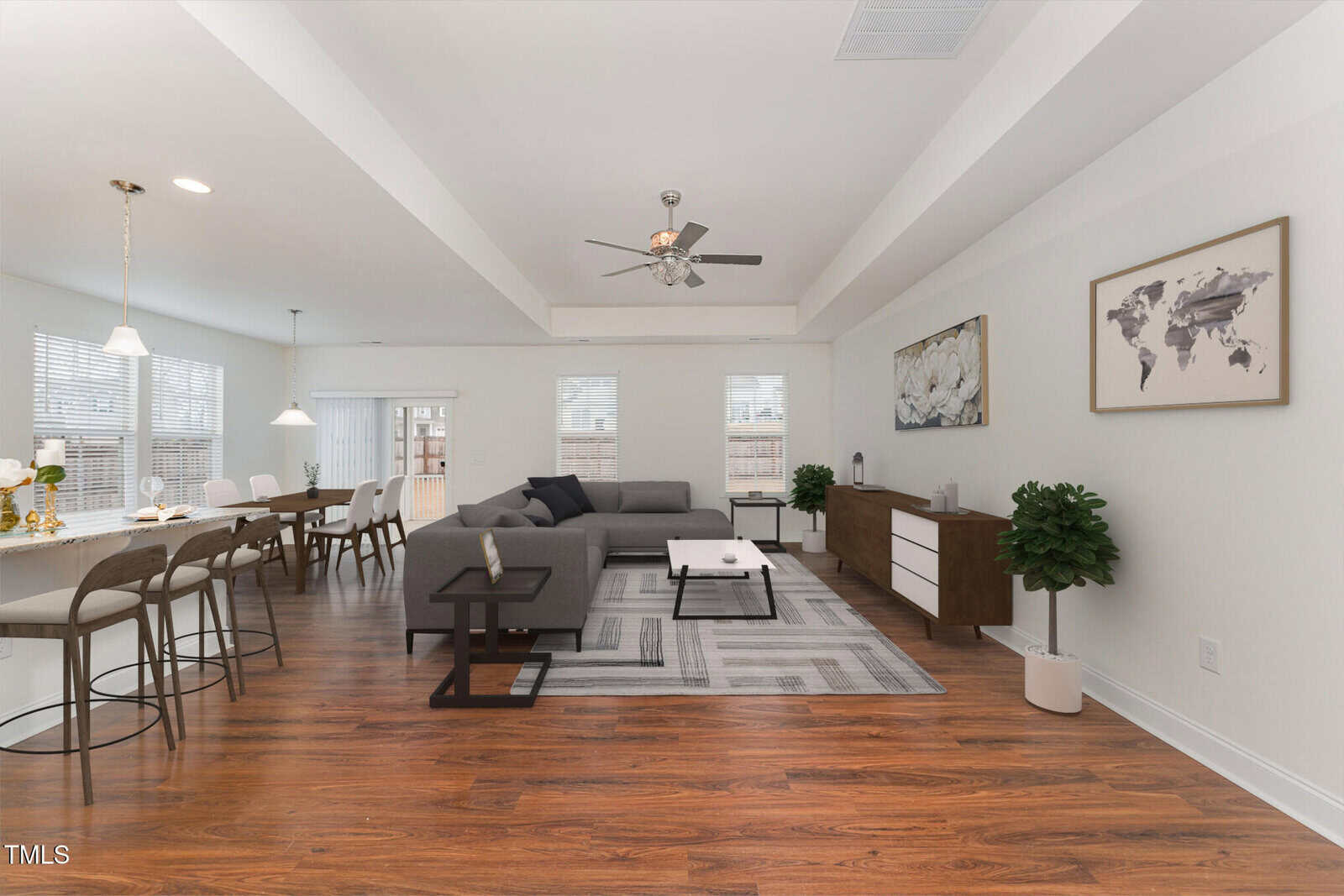 A modern open-concept living and dining area features wood flooring, a gray sectional sofa, a large rug, a ceiling fan, a dining table with chairs, a sideboard, two potted plants, and wall art.