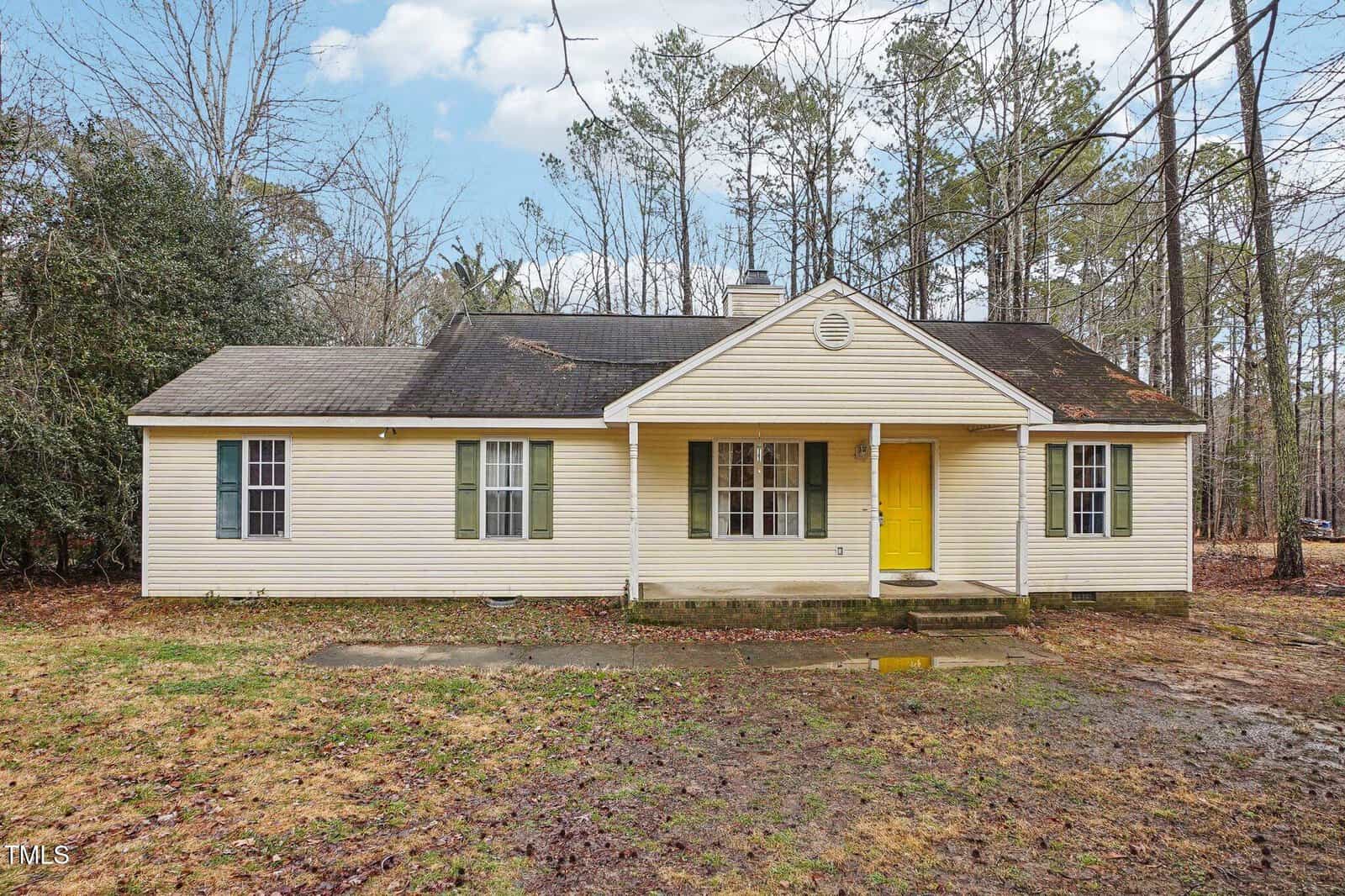 A single-story beige house with green shutters and a bright yellow front door, surrounded by trees and a patchy, muddy lawn under a partly cloudy sky.
