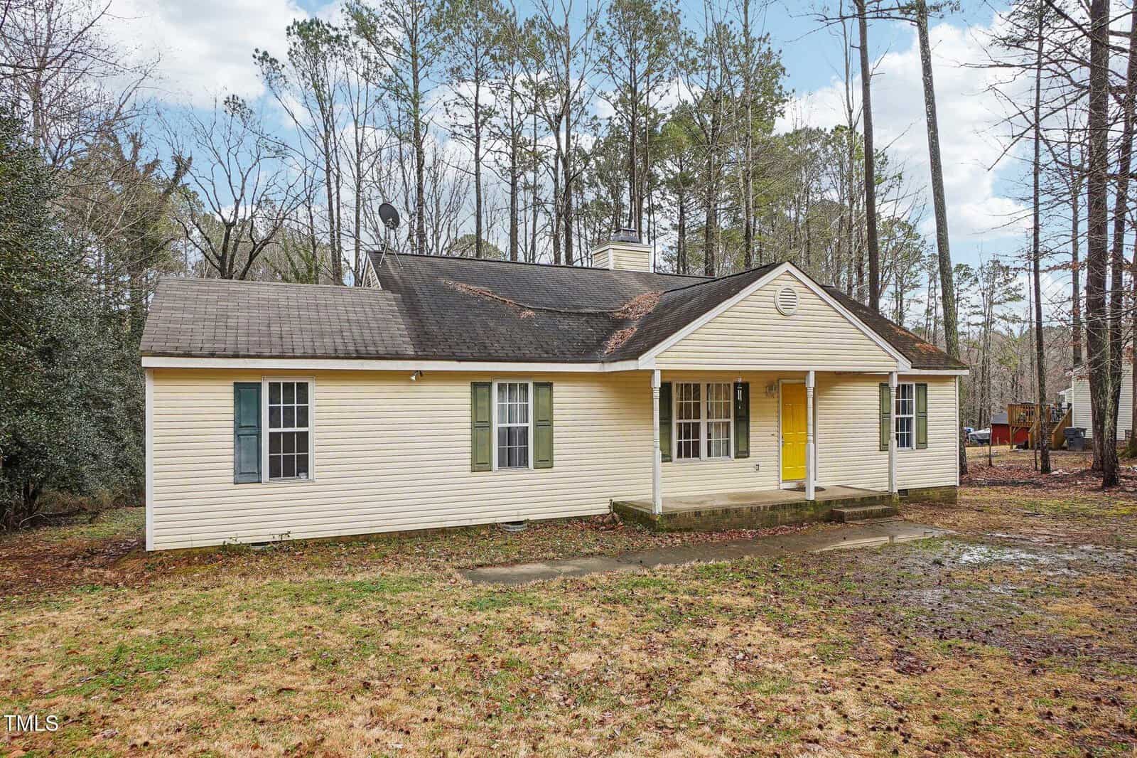 Single-story beige house with a dark roof, green and white shutters, a yellow front door, and a small porch. The house is surrounded by trees and sits on a patchy grass and dirt yard.