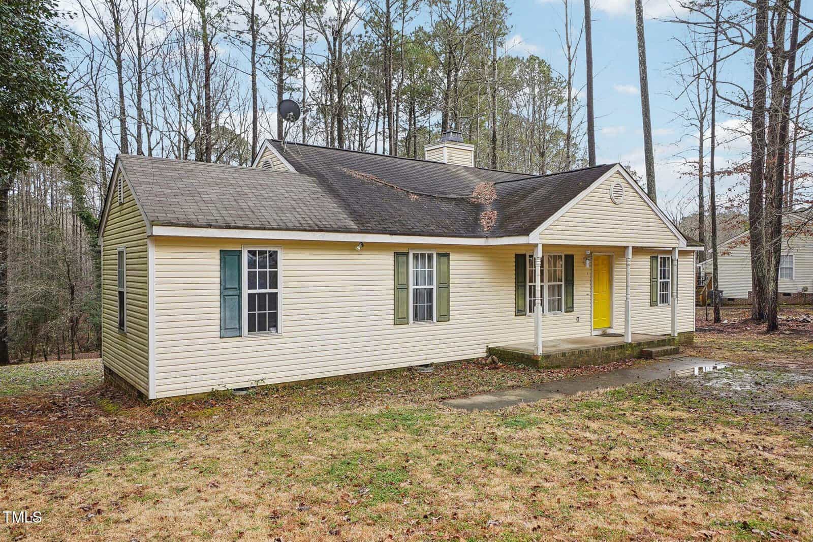 Single-story yellow house with green shutters and a yellow front door, small front porch with columns, surrounded by leafless trees and grass, overcast sky overhead.