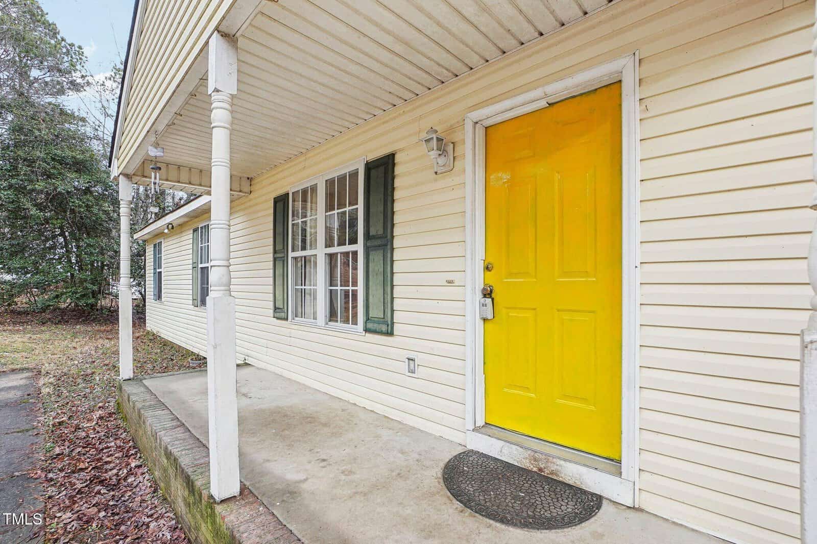 A house with yellow siding, a yellow front door, green window shutters, a small porch with white columns, a wall-mounted light, and a black doormat. Fallen leaves cover the ground beside the porch.