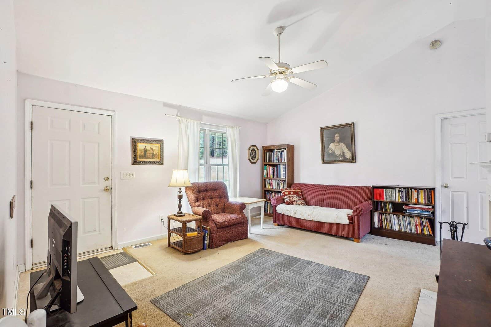 A living room with light walls, a ceiling fan, and carpeted floor. The space features a maroon armchair, matching sofa, bookshelves, a TV on a stand, framed artwork, a lamp, and a window with sheer curtains.