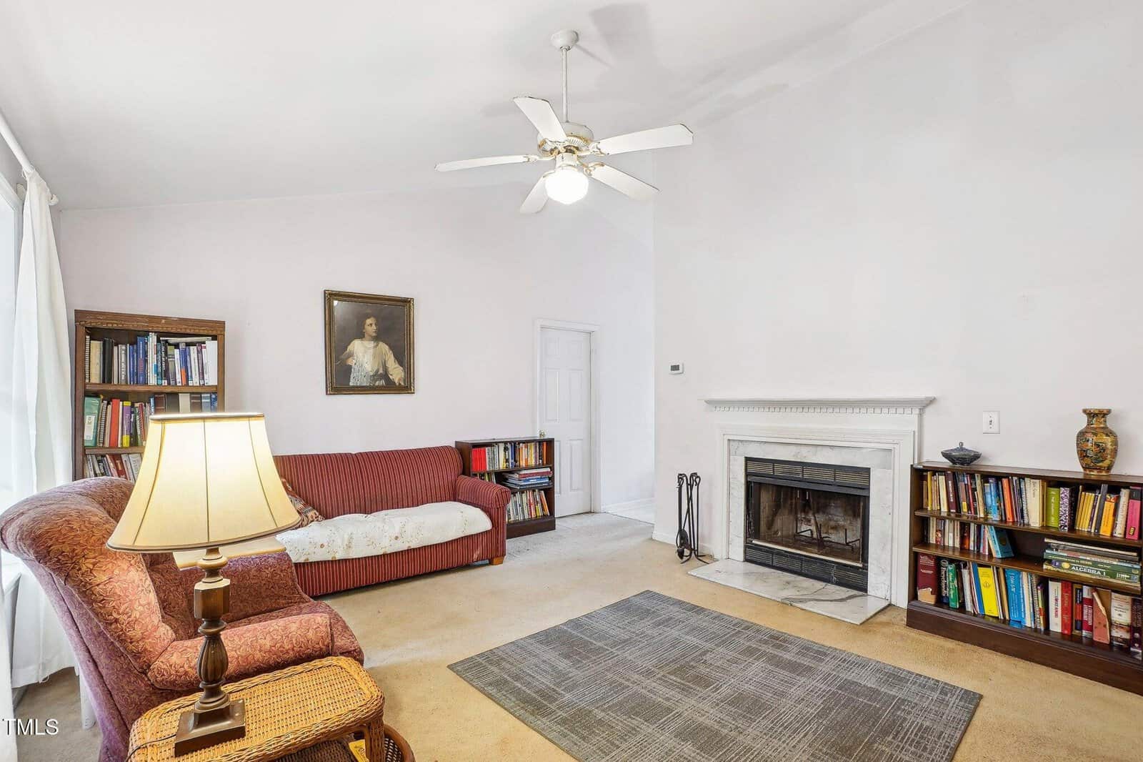 Living room with two bookshelves, a red striped sofa, a lamp, a fireplace with tools, a framed portrait above the sofa, and a ceiling fan. A rug is on the floor, and books and a vase are on the shelves.