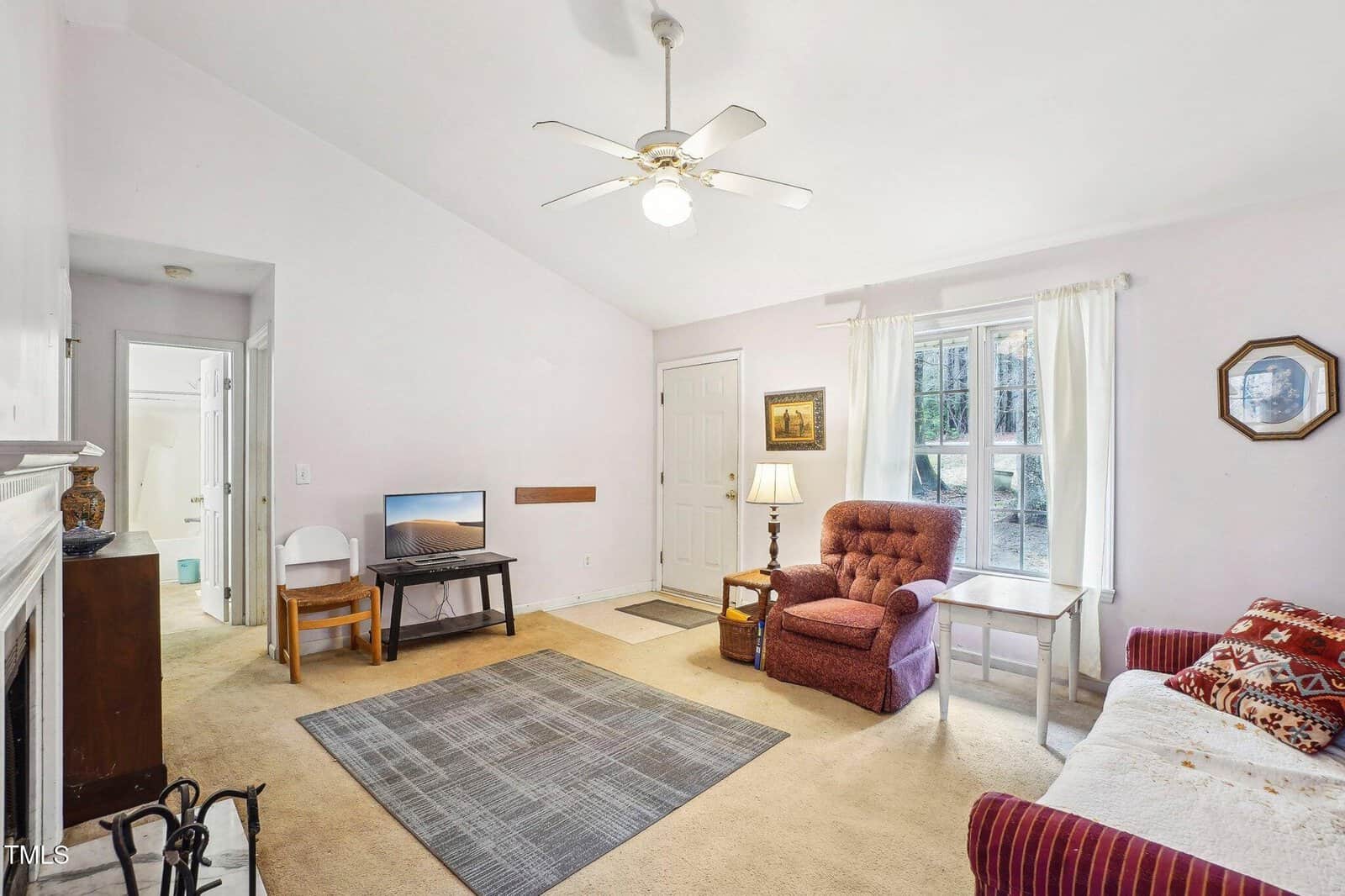Living room with beige carpet, area rug, armchair, side table, small TV on a stand, lamp, couch, wall art, and a ceiling fan. A window with white curtains and a door are visible. Bathroom entrance in the background.