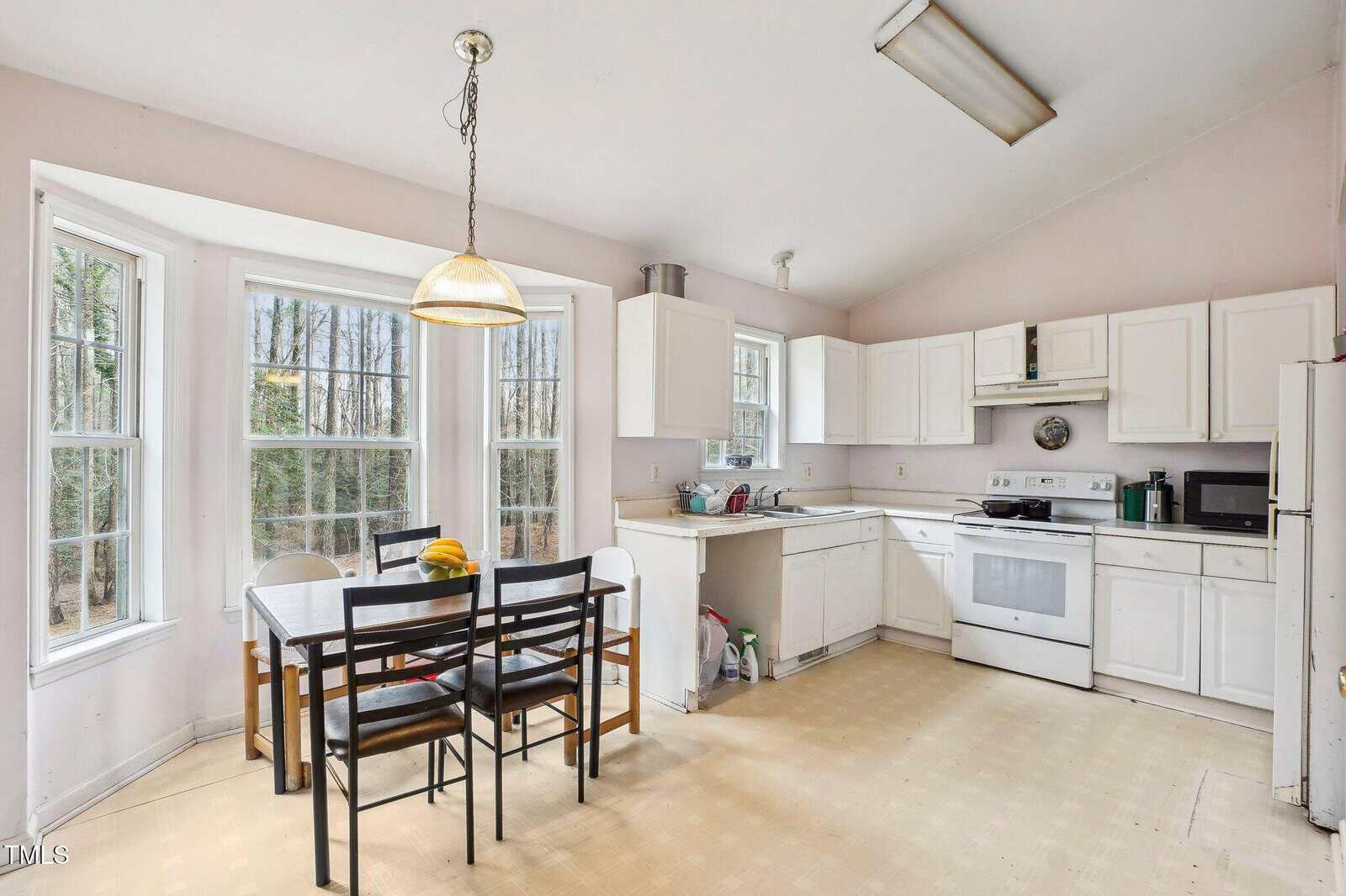 Bright kitchen with white cabinets, appliances, and a dining table with four chairs by bay windows. Light fixture hangs above the table. Trees are visible outside through the windows. Floor is light-colored linoleum.