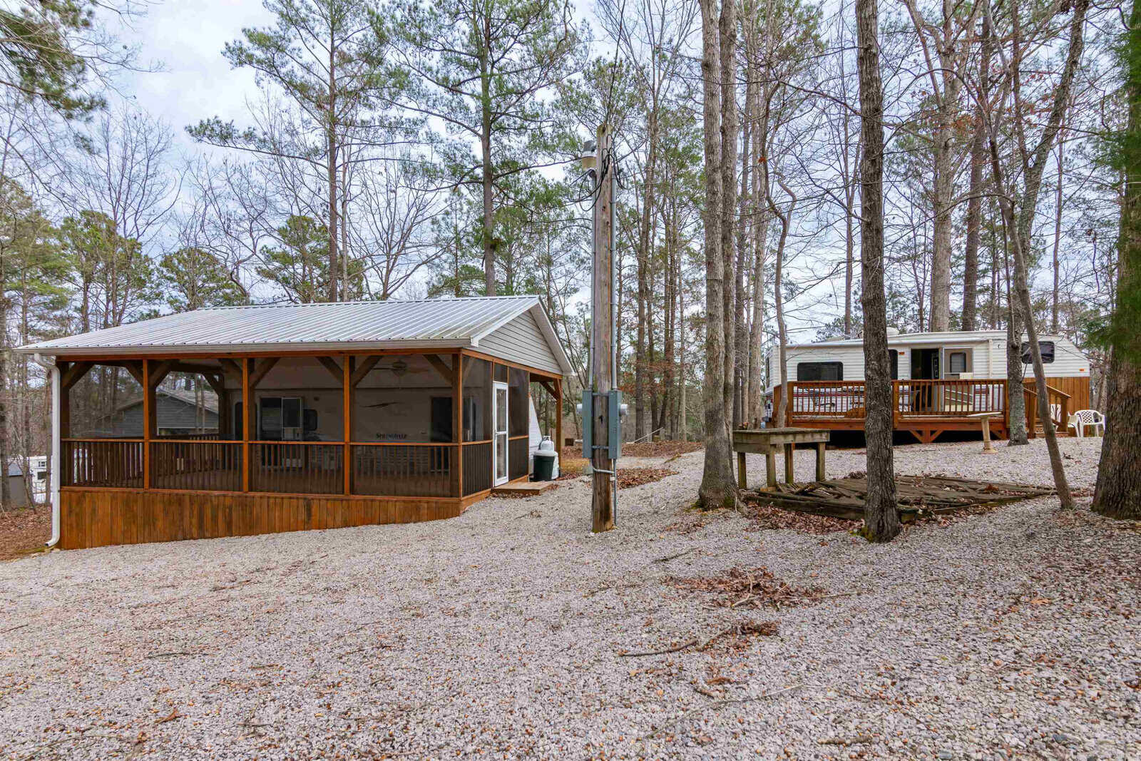 A small cottage with a screened porch sits on a gravel lot surrounded by trees, next to a travel trailer on a wooden deck. The area is quiet and wooded, with leafless trees and overcast skies.