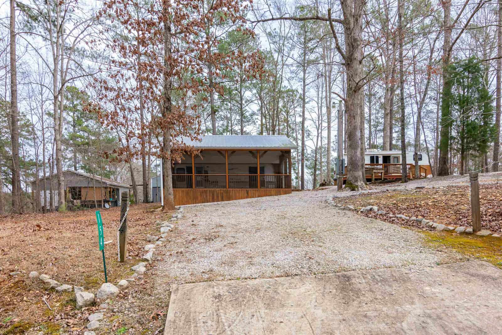 A gravel driveway leads to a wooden cabin surrounded by tall trees, with a covered porch in front. A small building and an RV are visible in the background among the trees. The scene appears calm and rustic.