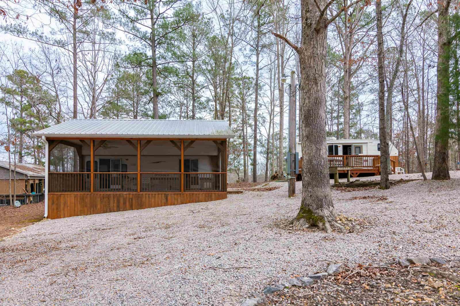A small wooden cabin with a covered porch and railing is set among tall trees on a gravel lot. In the background, a white RV is parked near the trees. The area appears calm and wooded.