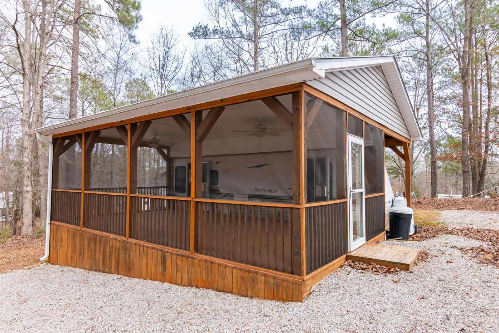 A small screened-in porch with wooden framing and a gravel pathway, attached to a single-story house surrounded by tall trees. A grill is visible on a small wooden platform near the entrance.