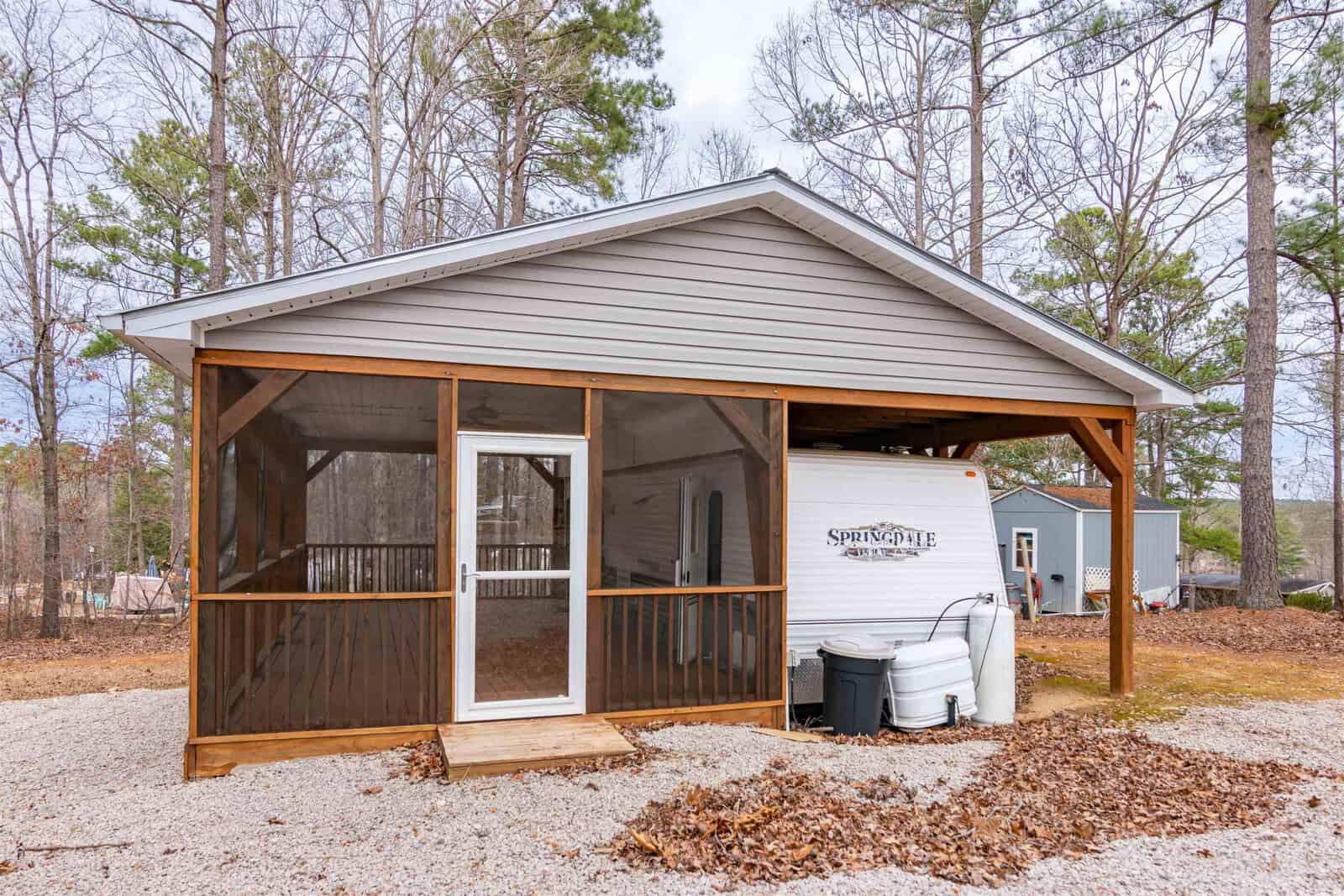 A small, screened-in wooden porch is attached to a simple gray carport structure, partially sheltering a white Springdale travel trailer. Gravel and dry leaves cover the ground, with trees and other buildings in the background.